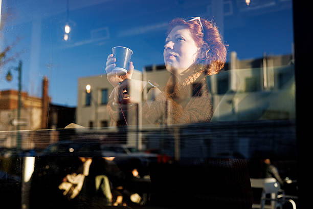 A 48-year-old Caucasian woman with red hair holds a paper cup while sitting inside a cafe. She is viewed through the window glass, which shows reflections of the outside buildings and street. The window creates a layered visual effect with the woman in focus while urban elements are reflected on the glass surface.