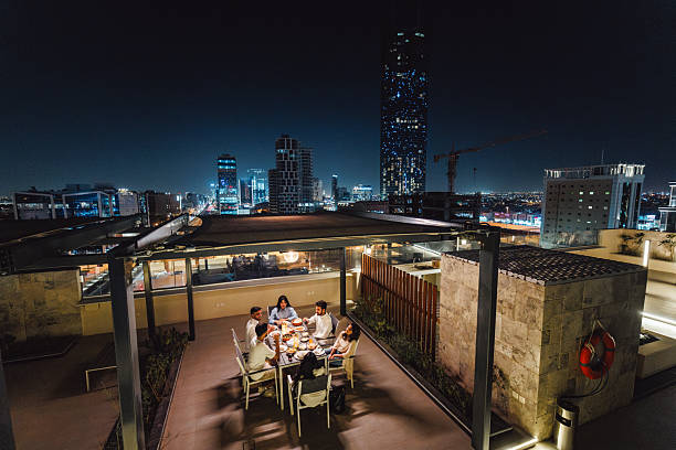 Group of cheerful young Arabic friends celebrating Ramadan on a rooftop terrace in Riyadh featuring a vibrant cityscape. They are dining together, engaging in lively conversation, and enjoying the festive atmosphere under the night sky.