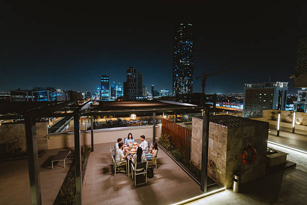 Group of cheerful young Arabic friends dining on a rooftop terrace in Riyadh featuring a vibrant cityscape at night. They enjoy a lively meal under the stars, surrounded by the illuminated skyline. Laughter and joy fill the air.