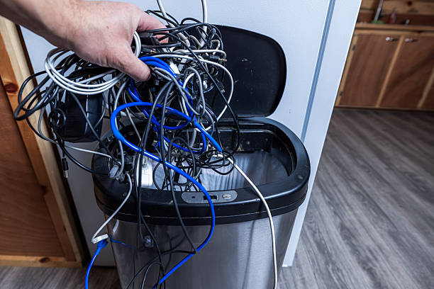 A high-angle conceptual shot of a hand discarding a large, tangled cluster of various electronic wires and cables into a stainless steel trash can.