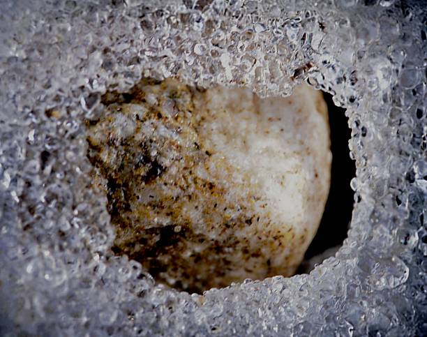 A masterfully balanced macro shot of a textured, light-toned stone peering through a natural opening in a thick sheet of ice. The subdued exposure reveals the intricate granular surface of the rock, contrasted against the cold, translucent geometry of frozen air bubbles