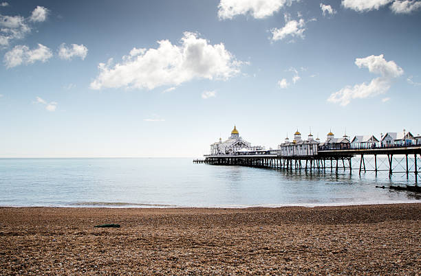 A historic Victorian pier stretches from a pebbled shore into tranquil blue waters beneath a bright, cloudy sky. Golden-domed buildings add charm as calm waves meet the coastline.