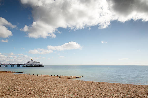 A tranquil coastal view featuring a pebble beach, wooden groynes, and a distant pier stretching into calm blue water under a bright sky with scattered clouds. Ideal for travel, leisure, and summer mood visuals.