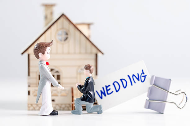 Two groom figurines, one kneeling to propose, stand before miniature house with a WEDDING card and binder clip on clean white background with copy space, expressing inclusive commitment and equality.