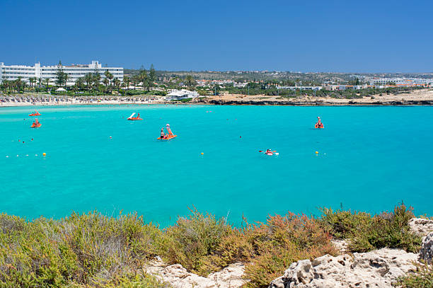 Wide panoramic view of a crowded Mediterranean beach with crystal clear turquoise water, swimmers, and people relaxing on the sandy shore. Luxury resort area with palm trees and hotels in the background under a clear blue sky. Concept of summer vacation, tourism, seaside leisure, travel destination, coastal lifestyle, holidays, and warm climate. Ideal for travel agencies, tourism promotion, resort marketing, summer advertising, and lifestyle content.
