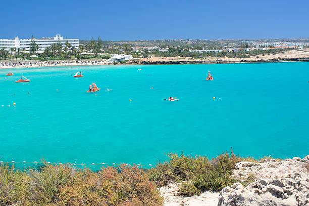 Wide panoramic view of a crowded Mediterranean beach with crystal clear turquoise water, swimmers, and people relaxing on the sandy shore. Luxury resort area with palm trees and hotels in the background under a clear blue sky. Concept of summer vacation, tourism, seaside leisure, travel destination, coastal lifestyle, holidays, and warm climate. Ideal for travel agencies, tourism promotion, resort marketing, summer advertising, and lifestyle content.