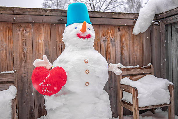 Funny handmade snowman holding a red heart in a snowy backyard. Winter love and Valentine concept.