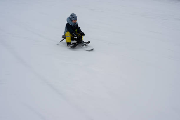 Young child wearing a hat and warm clothing sledding down a snowy hill on a ski sled, enjoying winter sports outdoors.