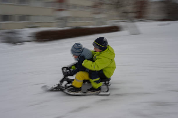 Happy little boy dressed in a green winter jacket, blue pants, black boots, and a red‑and‑black knit hat speeds downhill on a bright yellow sled in a snow‑covered forest. The background trees and snow are captured with intentional motion blur, emphasizing the sense of speed and excitement. Snow trails behind the sled add to the dynamic energy of the scene. The image conveys the thrill of sledding, childhood joy, and the beauty of outdoor winter recreation.Shot with Nikon D800