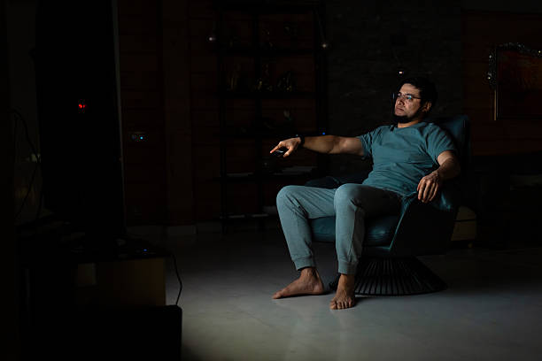 Man in armchair pointing remote at tv in a dark living room at night, relaxing and watching television alone