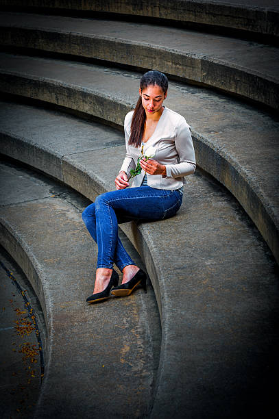 Woman Sitting on Stone Steps. A woman sits on curved stone steps holding a rose. She is dressed in casual attire with blue jeans and a light jacket.