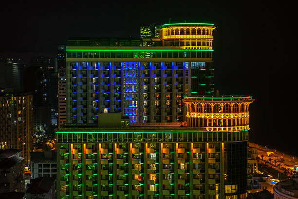 A neon lit high rise in Batumi, Georgia, shows arched colonnades, terraces, twin rooftop cylinders, and many balconies, set against a dark sky and dimmer nearby buildings.