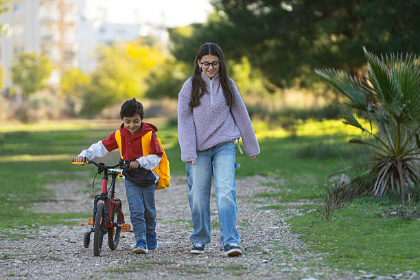 A sister walks next to her younger brother as he rides a bicycle while they are going to school together.