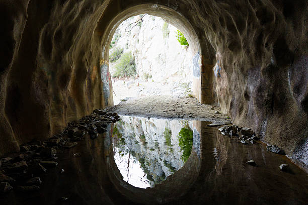Bright, rocky entrance of an abandoned tunnel is perfectly reflected in the still water of a puddle on the floor, creating a stunning symmetrical image.