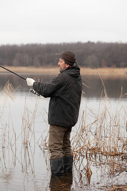 A man in white gloves fishes with a fishing rod with a reel. A fisherman in rubber boots stands in the middle of a lake in the water. Reeds grow around