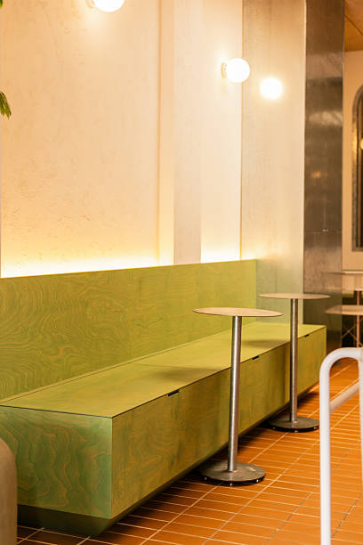 In a modern cafe, two small round tables stand beside a long green bench. The area is well-lit with warm light, creating a welcoming atmosphere for visitors