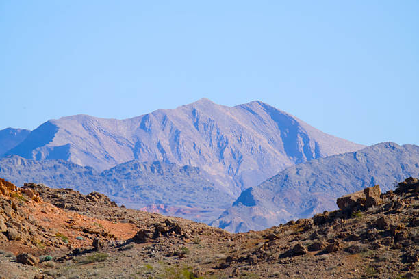 A distant desert mountain ridge rises beyond a rocky foreground in southern Nevada, showing layered terrain and arid conditions under a clear sky.