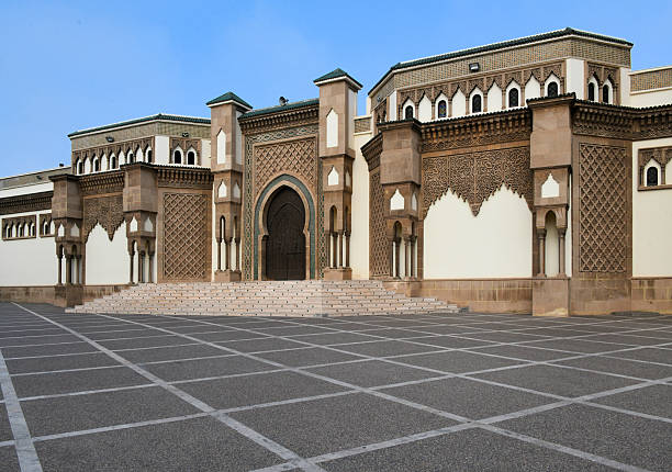Agadir, Souss-Massa, Morocco: exterior view of the Mohamed V Mosque, located in the Talborjt district. The image displays the main entrance and facade, characterized by traditional Moroccan-Andalusian architectural elements. The structure features intricate carved stonework, geometric wood carvings, and zellij tilework. Named after King Mohamed V, who led Morocco to independence, the mosque serves as a significant religious and political landmark in the city, which was largely rebuilt following the 1960 earthquake. The temple adheres to the Maliki school of Sunni Islam, reflecting the regional aesthetic through horseshoe arches and decorative motifs. The foreground consists of a large, paved public plaza.