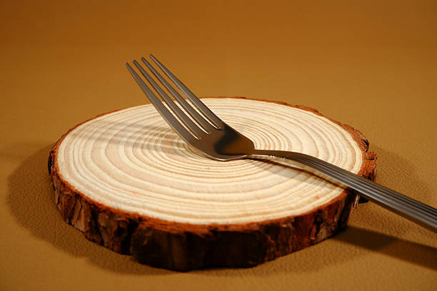 Shiny metal fork dramatically positioned atop a rough-cut circular wood slab. This closeup highlights the reflective surfaces and the contrast between modern stainless steel and natural wood grain presentation elements.