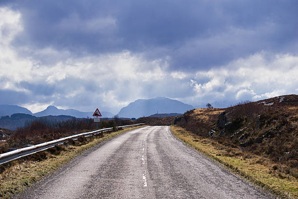 Atmospheric landscape photograph of an empty road along Scotland’s NC500, leading toward imposing Highland mountains in the distance. Autumn colors cover the landscape, while a richly textured, cloud-filled sky adds drama and depth. Perfect for travel, road trip, and scenic landscape stock imagery.