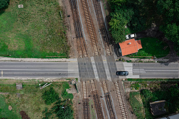Aerial image of a railway crossing intersecting a two-lane road, with a car passing over the tracks. A small red-roofed building and surrounding greenery add contrast to the geometric layout of the tracks and road. The composition blends transportation infrastructure with rural simplicity.