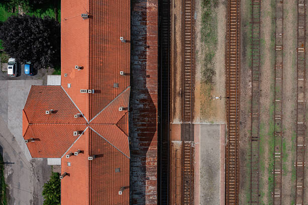 Top-down aerial photograph of a vintage train station with red tiled roofs next to multiple rusty and overgrown railway tracks. The geometric symmetry of the roof contrasts with the parallel lines of the tracks, creating a visually striking industrial composition. The muted tones and weathered textures evoke a sense of nostalgia and abandonment.