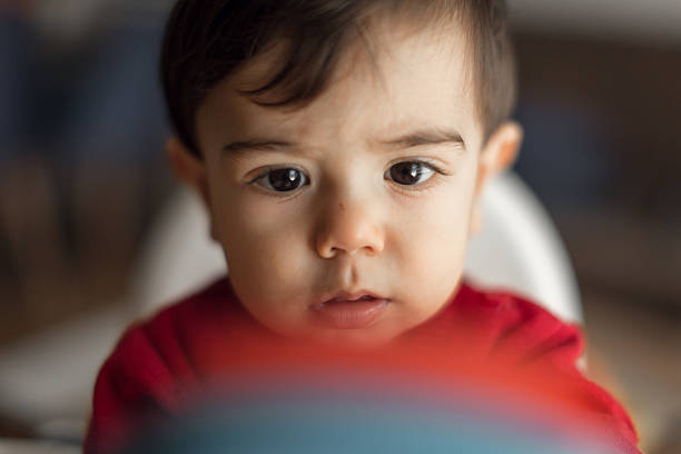 Close-up shot of a cute baby in a red shirt, focusing intently. The background is softly blurred, highlighting the baby's facial expression and the vibrant colors in the foreground.