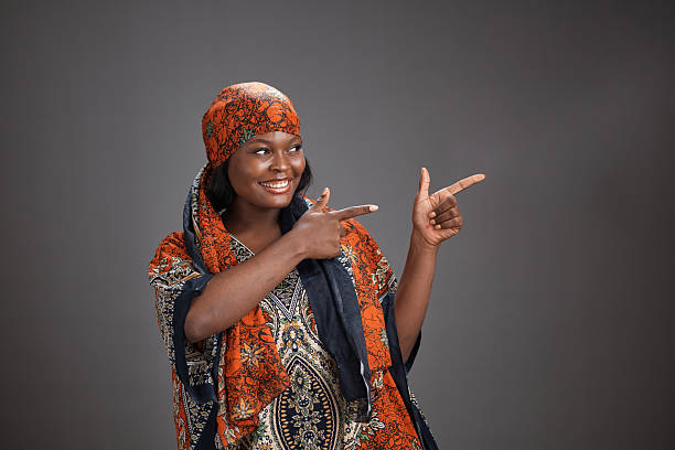 Full-length portrait of a beautiful young African girl dressed in traditional clothing and a headscarf standing against a plain brown yellow warm background listening to music through headphones  on her mobile phone in Dar es Salaam East Africa