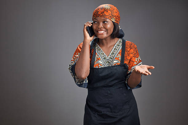 Young African woman wearing a patterned traditional dress, headscarf, and apron smiles while talking on a smartphone, gesturing with an open palm against a plain gray studio background with copy space.