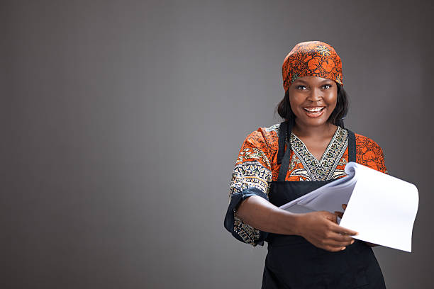 Studio portrait of a smiling woman wearing traditional patterned clothing and headscarf while holding papers, standing against a grey background with copy space.