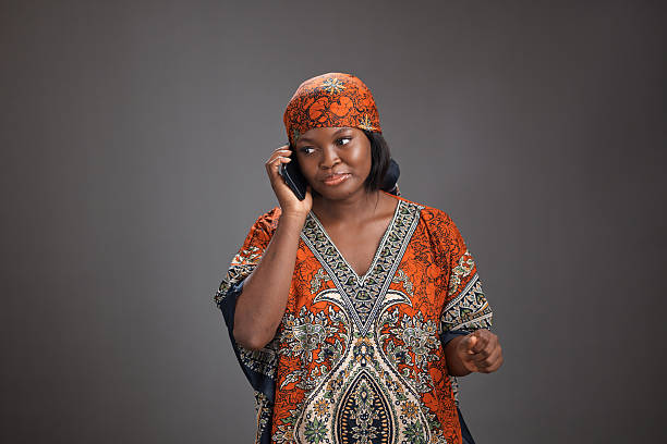 Young black woman wearing a patterned traditional dress and matching headscarf listens while talking on a smartphone, standing in a studio against a plain gray background with soft lighting.