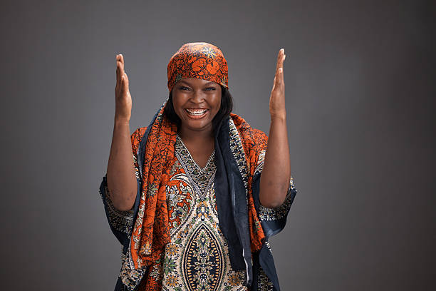 Young African woman wearing a patterned traditional dress and matching headscarf smiles with raised hands and an open mouth, standing against a plain gray studio background while expressing joy and excitement.