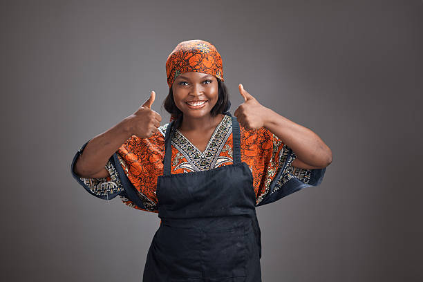 Young African woman wearing a patterned traditional dress, headscarf, and apron smiles while giving a thumbs up with both hands, standing against a plain gray studio background with a positive expression.