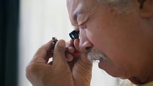 Elderly Asian man with long white hair and a mustache examines his collection of sacred Buddhist amulets while referencing information on a tablet, sipping coffee during a peaceful morning at home. A calm, wellness-inspired scene that highlights culture, mindfulness, and personal hobbies.
