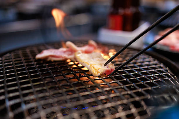 Close-up view of thin slices of wagyu beef being grilled over an open flame on a metal barbecue grill, held with chopsticks. The image highlights sizzling meat, fire, and authentic Asian barbecue dining experience, with selective focus.