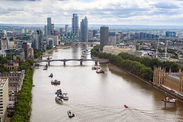 A sweeping aerial view of Westminster, the Houses of Parliament, Big Ben, and the Thames in central London. Boats dot the river while the city stretches into a dense, green-bordered skyline.