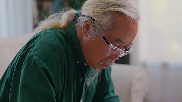 Elderly Asian man studies through an online class on his smartphone while carefully taking notes in a notebook. The scene reflects a wellness-focused senior lifestyle, highlighting lifelong learning, digital literacy, and calm concentration in a comfortable home environment.