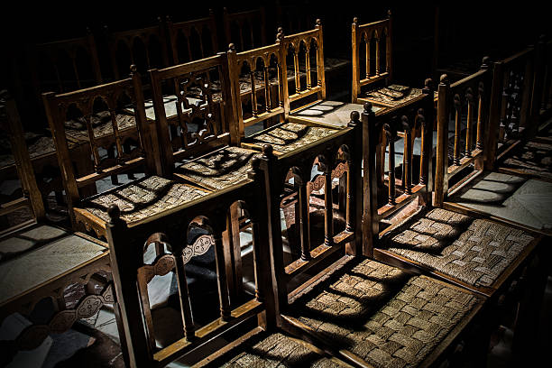 Empty chairs in Monreale cathedral, Sicily, Italy. The woven straw seats are a rustic contrast to the refined woodwork of the backrests. Neat repeating rows create a sense of order & rhythm. Chiaroscuro, an interplay between light & shadow. The scene is plunged into a deep velvety darkness from which the chairs emerge. Luminous areas are juxtaposed with deep impenetrable shadow. An atmosphere of stillness, quiet solitude, audible silence
