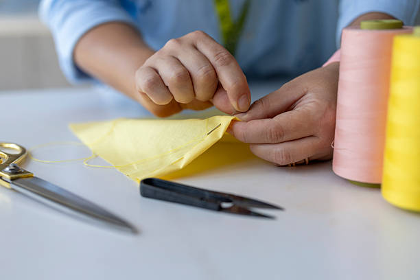 Detailed close-up view of a seamstress or tailor's hands stitching a piece of yellow cloth using a needle and thread. The workspace is arranged with sewing accessories including gold scissors, colorful thread spools, and snips, representing fashion design, DIY crafts, and clothing repair.