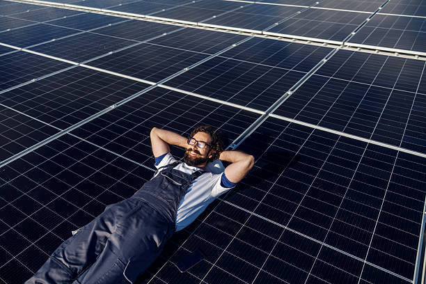 Male worker lying on a solar panel array, resting with hands behind his head and eyes closed, enjoying the warm sunlight after completing work on a sustainable energy project