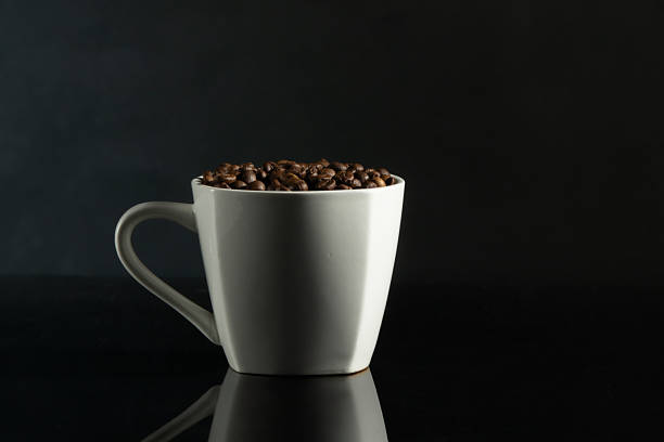 Minimalist still life of a white ceramic coffee cup filled with roasted coffee beans on a dark black background with reflection. Clean modern composition with copy space, representing coffee, aroma, cafe