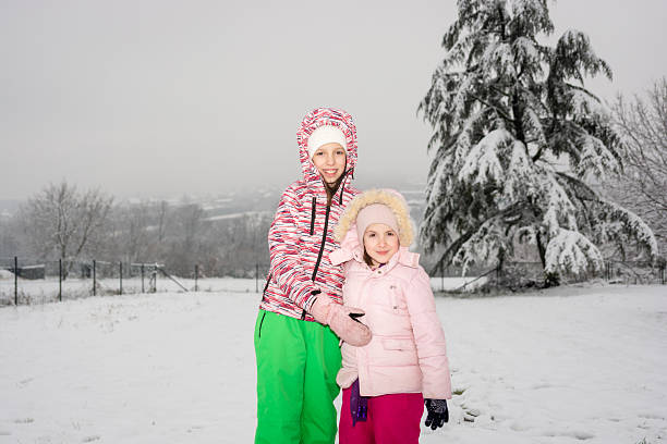 Two cheerful young sisters wearing warm winter clothing standing in fresh snow on a cold day, smiling at the viewer