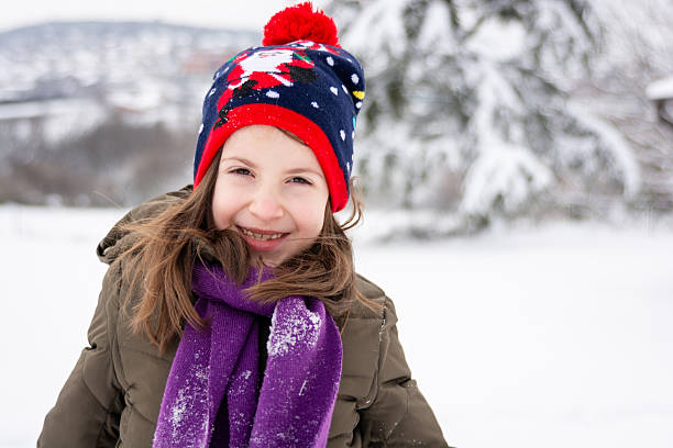 Cheerful elementary age girl smiling at camera, wearing a knit hat and purple scarf in a snowy winter landscape