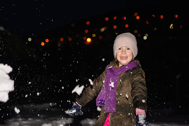 Smiling elementary-age girl playing joyfully in falling snow at night, bundled in warm winter clothes for holiday fun