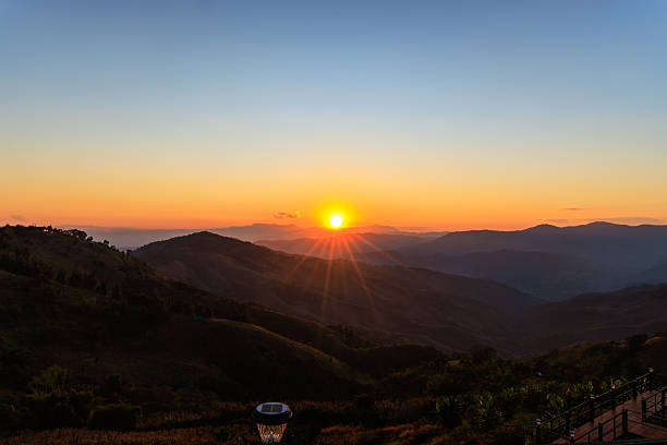 A stunning sunset illuminates mountain peaks with distinct golden sun rays. The scenic landscape features rolling hills and a wooden viewing deck in the foreground. A peaceful evening atmosphere.