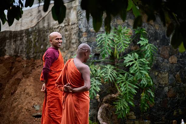 Two monks standing near stone wall and foliage.