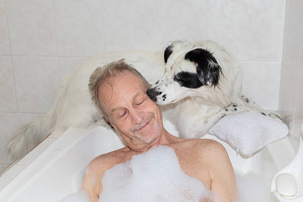 A senior man relaxes in a bubble bath while his dog gently nuzzles his face in a bright home bathroom. The image captures a tender everyday moment between a pet and owner, highlighting companionship, trust, emotional support, and the rewarding bond of pet parenting.