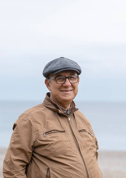 Portrait of a kind, smiling elderly man with glasses and a stylish flat cap, standing against a soft-focus background of the sea. This image represents a peaceful retirement, healthy aging, and active senior lifestyle