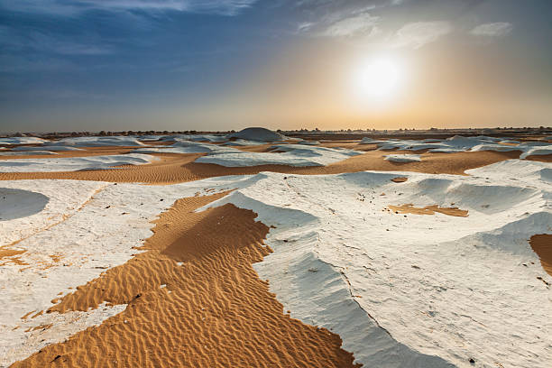 A wide, otherworldly landscape of pale, chalk-like rock formations interspersed with warm orange sand dunes. The low sun casts golden light and soft shadows across wind-sculpted ridges and rippled sand, highlighting contrasting textures and shallow hollows that stretch toward a distant horizon beneath a clear, gradient sky.
