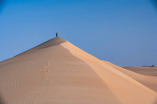 A solitary person stands with arms raised on the crest of a large sand dune, silhouetted against a clear blue sky. A single line of footprints winds up the dune's ridge through rippled, golden sand. Soft side lighting casts gentle shadows that emphasize the dune's curves, conveying a sense of scale, isolation, and triumph.